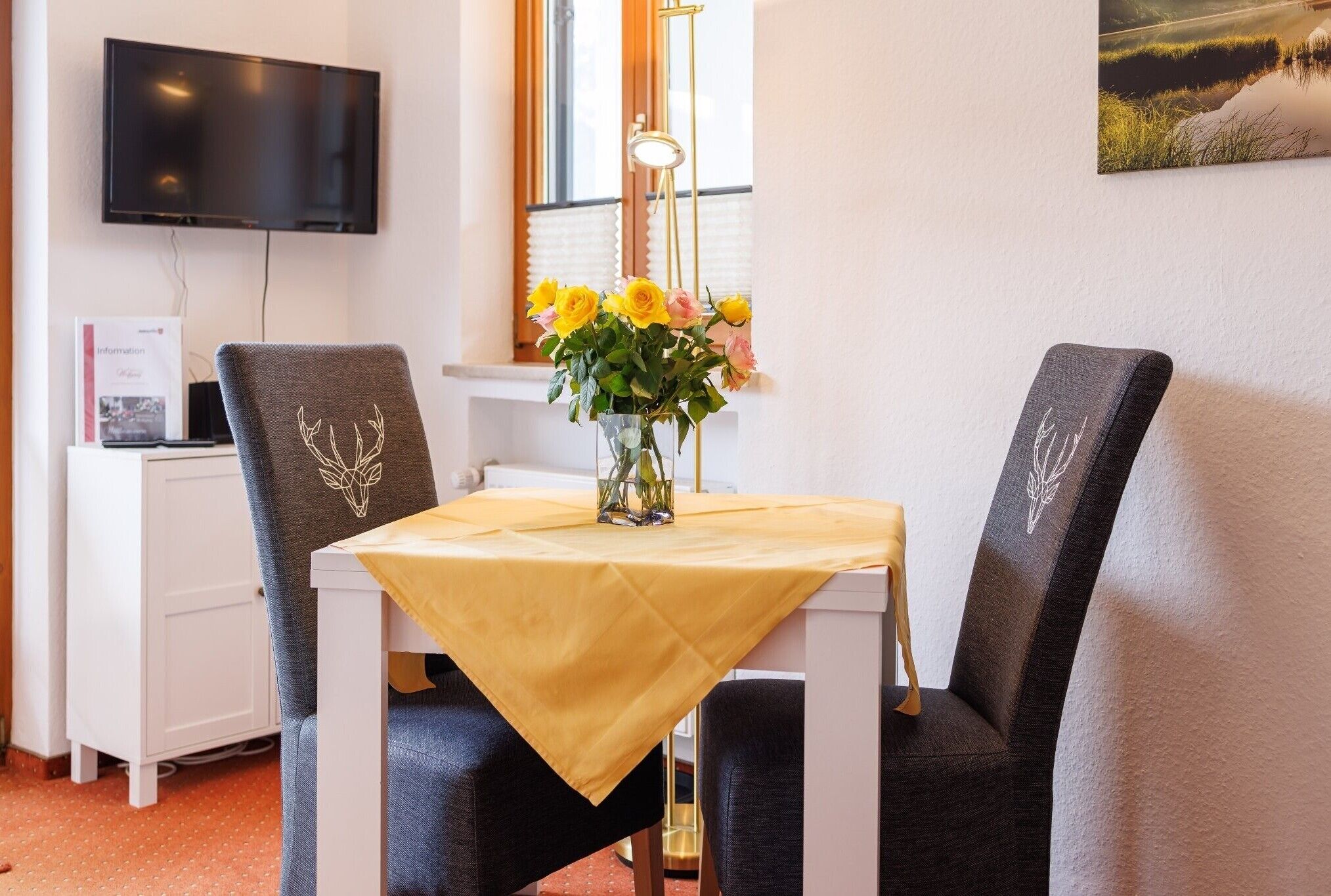 Ein Essbereich mit Tisch für zwei Personen, einem Blumenstrauß, einem Fernseher und einem Fenster mit Jalousien.