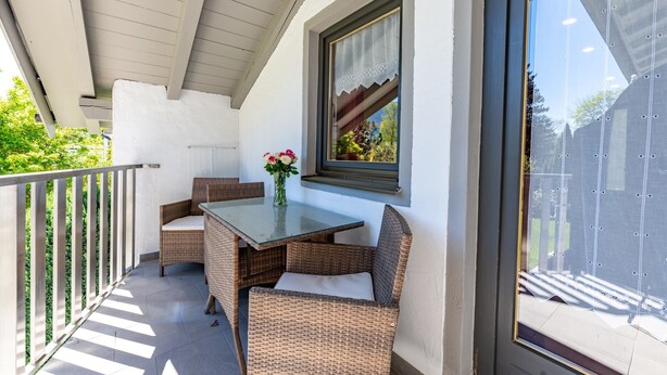 Ein Balkon mit Korbmöbeln, einem Glastisch und einem Fenster mit Blick auf Grünflächen.