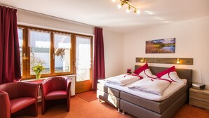 A hotel room with a bed, two chairs, a desk, a window with curtains, a painting on the wall, and a nightstand.
