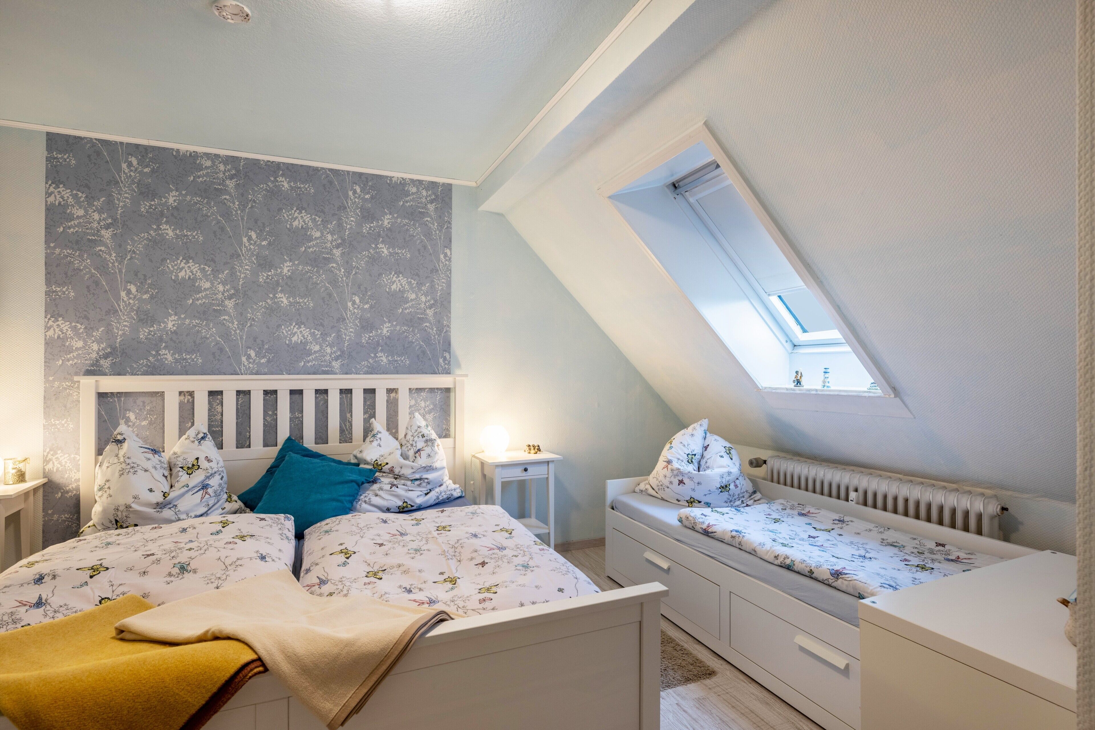 Une chambre avec un plafond en pente, un puits de lumière, un lit avec une literie à motifs floraux, une table de chevet et un radiateur.