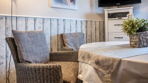 Una habitación con una silla de mimbre, una mesa con mantel, un televisor sobre un mueble y un cuadro en la pared.