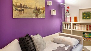 A room with a purple wall, a painting of a harbor, a bookshelf, and a bed with pillows. - Holiday Apartment 'Lila' with Shared Garden and Private Beach Chair (Nordstrand)