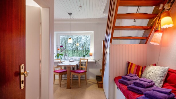 A room with a red sofa, a dining table with chairs, a window with a view of trees, and a wooden ceiling. - Holiday Apartment 'Lila' with Shared Garden and Private Beach Chair (Nordstrand)