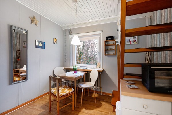 Une pièce avec une table en bois, deux chaises, un miroir, une étagère remplie de livres, un téléviseur et une fenêtre donnant sur des arbres.