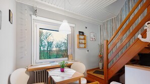 A dining area with a table, chairs, a hanging light, a window with a view, and a wooden staircase. - Holiday Apartment 'Op'nböhn' with Shared Garden and Private Beach Chair (Nordstrand)