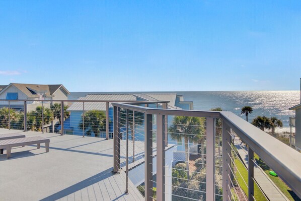 A balcony with a view of the ocean, a pool, and a building with a sloped roof.