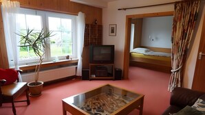 Een kamer met een televisie, een glazen tafel met een model, een bankje, een bank en een slaapkamer met gordijnen, plus een stapelbed.