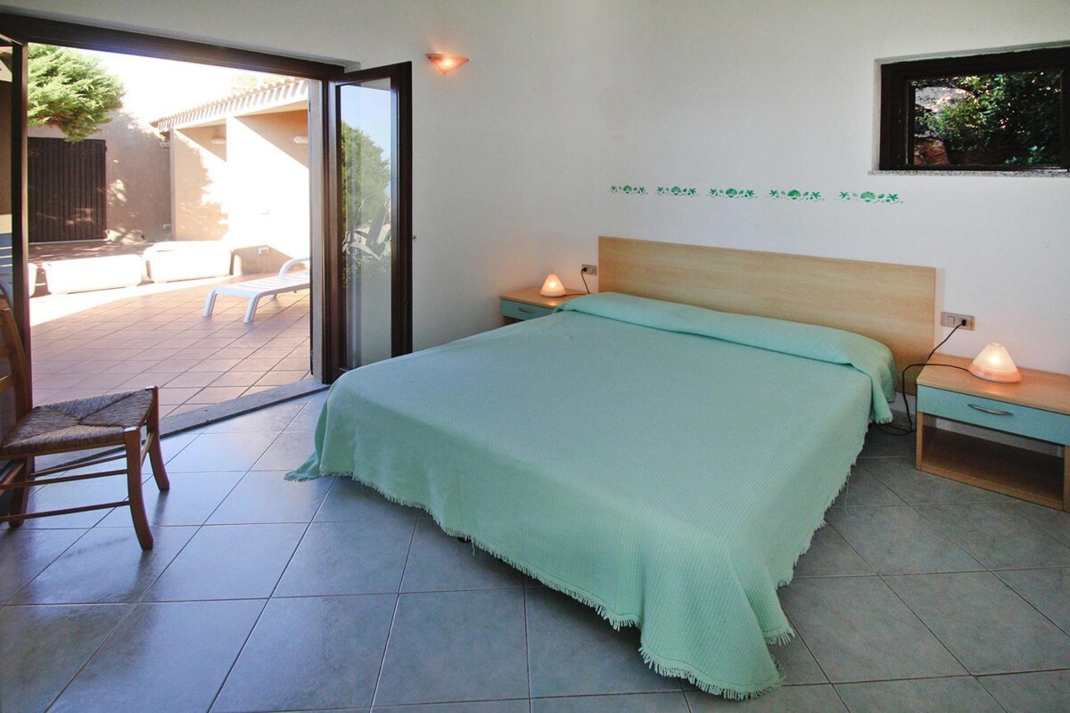 Une chambre à coucher comprenant un lit, deux tables de chevet, une chaise et une porte-fenêtre coulissante donnant sur un espace extérieur.