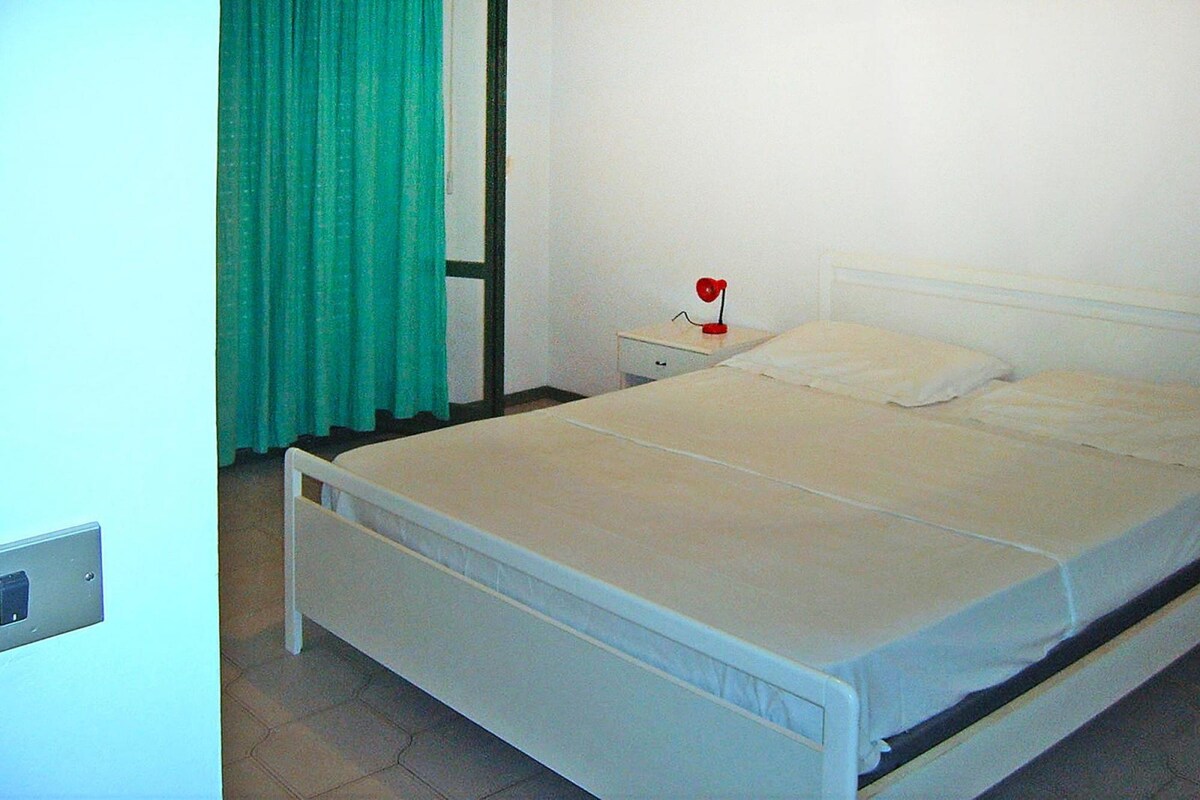 Une chambre avec un lit simple, un lit blanc, une table de chevet avec une lampe rouge, un rideau vert et un mur blanc.