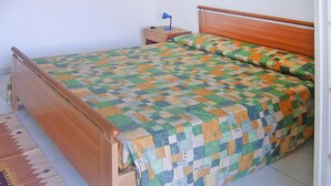 Sängyssä on ruutukuviollinen päiväpeite, puinen pääty, yöpöytä, jossa on lamppu, ja kuviollinen matto.