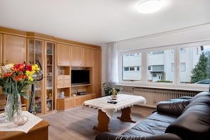 A living room with a sofa, a table with a lace tablecloth, a TV, wooden cabinets, and a window with a view of buildings.