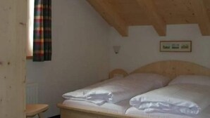 Una habitación con techo de madera, una cama con ropa blanca, un taburete pequeño y una lámpara fijada a la pared.