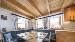 Un comedor acogedor con una mesa de madera y bancos tapizados en azul, una cocina con armarios de madera y ventanas amplias con cortinas.