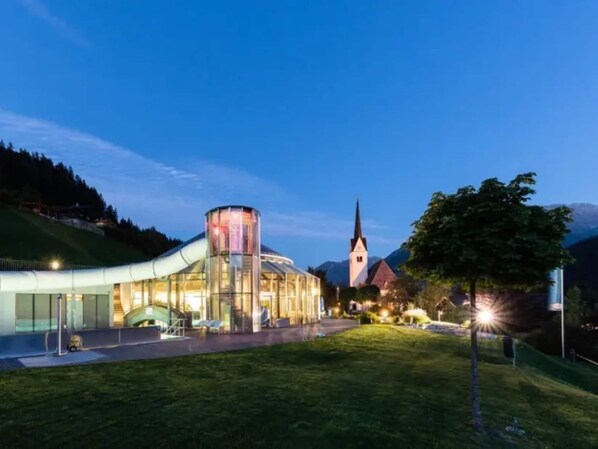 A modern building with a glass facade and a cylindrical tower, set against a backdrop of a church and mountains.