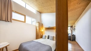 Un dormitorio con techo de madera, una cama con manta gris, una mesita de noche y una ventana con cortinas.
