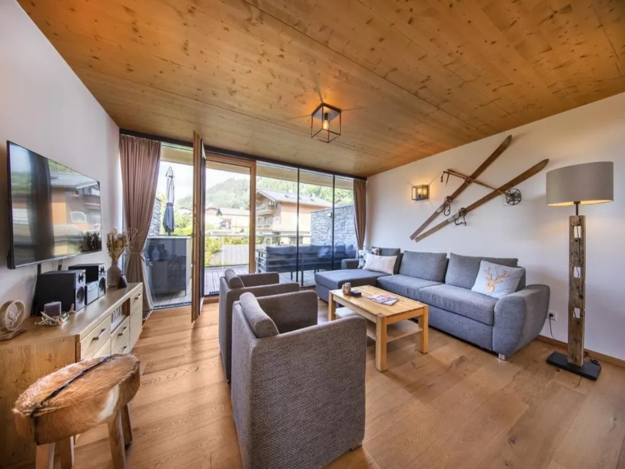 A modern living room with a wooden ceiling, a flat-screen TV, a grey sofa, a coffee table, and a large window offering a view of a mountain.