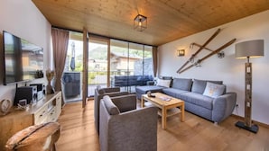 Un salón moderno con techo de madera, un televisor de pantalla plana, un sofá gris, una mesa de centro y un amplio ventanal con vistas a la montaña.