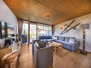 A modern living room with a wooden ceiling, a flat-screen TV, a grey sofa, a coffee table, and a large window offering a view of a mountain.