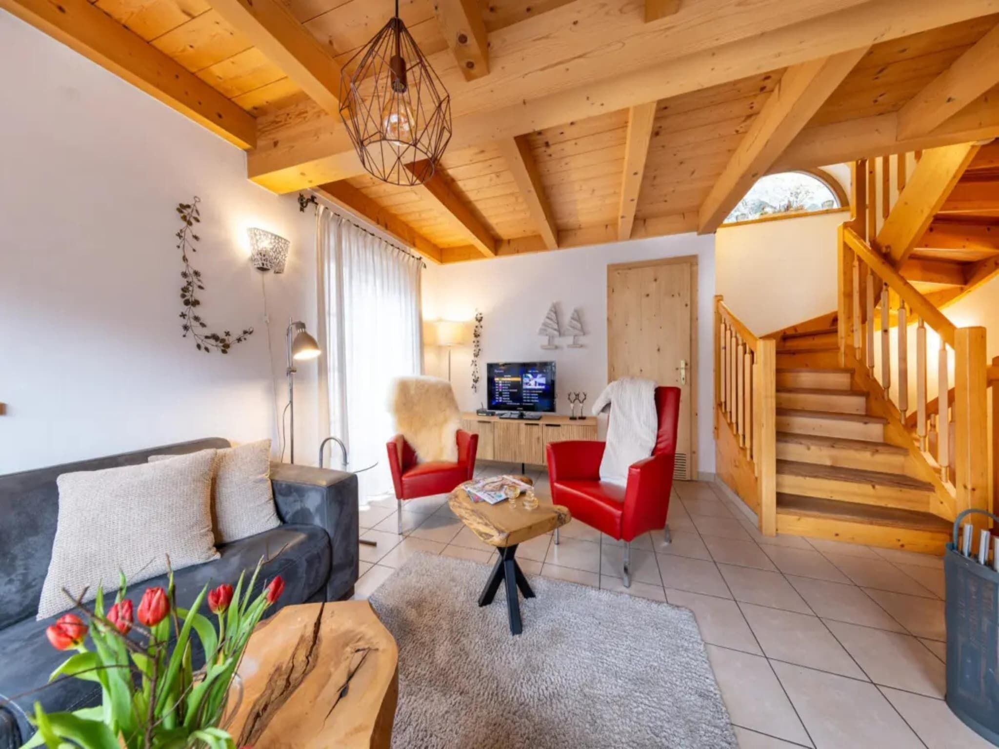 Ein Wohnzimmer mit einer Couch, zwei roten Sesseln, einem Couchtisch und einem Fernseher. Eine Holztreppe befindet sich rechts.