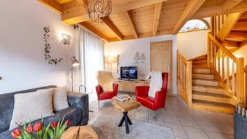 Ein Wohnzimmer mit einer Couch, zwei roten Sesseln, einem Couchtisch und einem Fernseher. Eine Holztreppe befindet sich rechts.