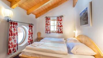Ein Schlafzimmer mit Holzboden, einem Bett mit weißer Bettwäsche, einem runden Fenster und roten, blumigen Vorhängen.