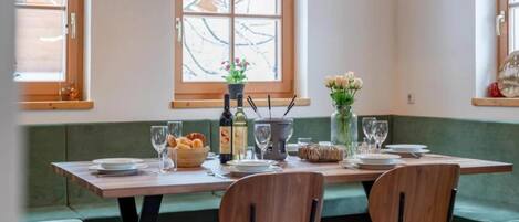 Un comedor con mesa de madera dispuesta para dos, un florero con flores y un ventanal con vista a árboles desnudados.