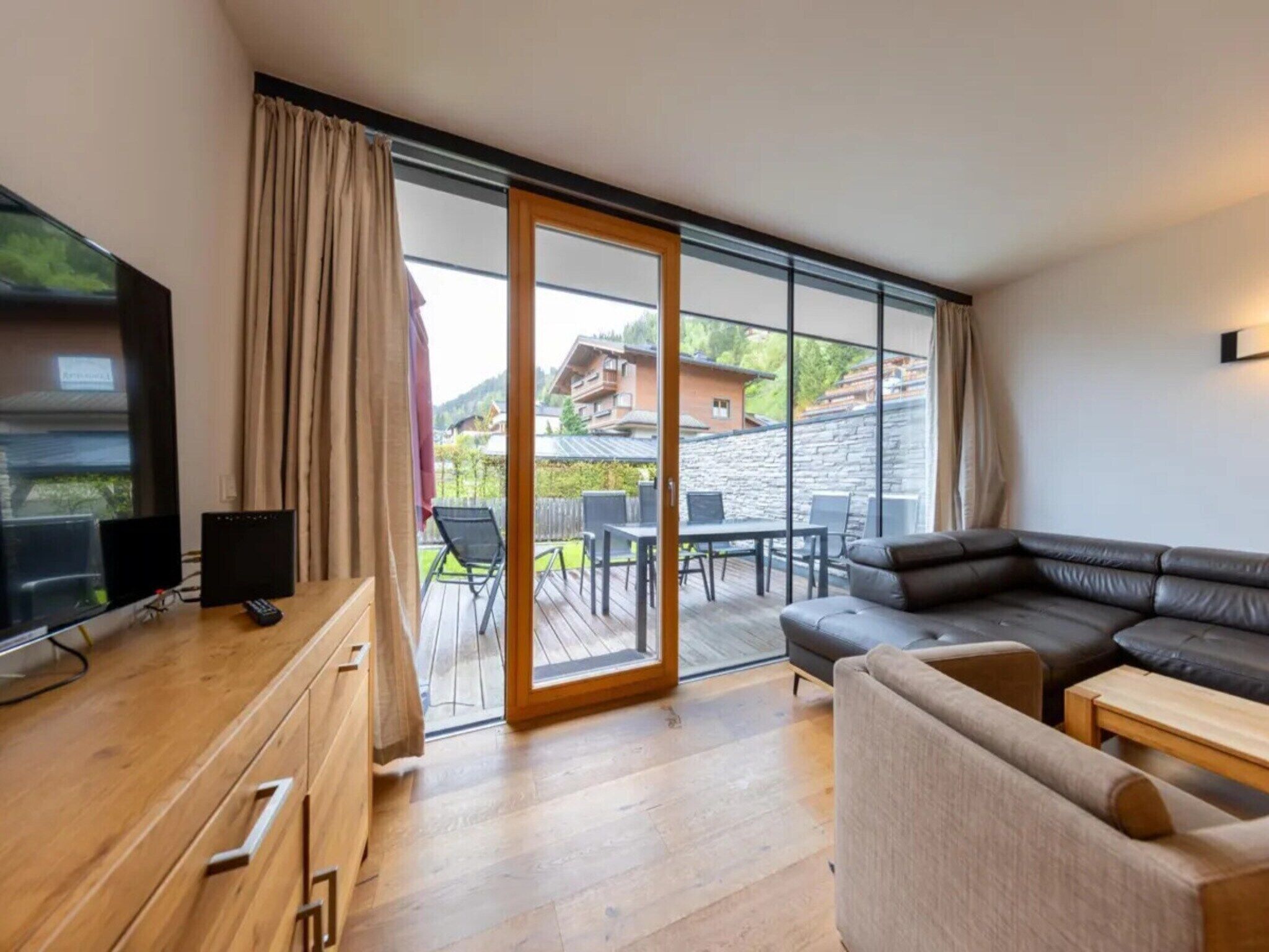 Un salon moderne comprenant un bureau en bois, un téléviseur, un canapé et une porte-fenêtre coulissante donnant sur une terrasse extérieure.