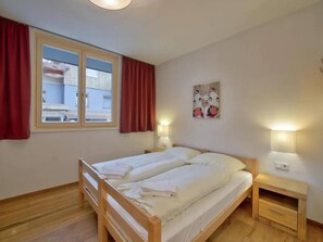 A bedroom with a double bed, two bedside tables, a window with curtains, and a wall-mounted light fixture.