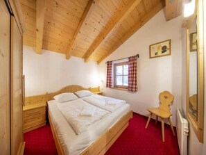 A bedroom with a wooden ceiling, a bed, a chair, a window with curtains, and a radiator.