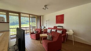 A spacious living room with a large window, a red sofa set, a television, and a white side table. - Zirbenholz Alpine Maisonette Stay (Wald-Königsleiten)