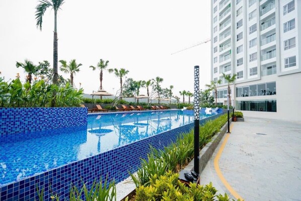 A swimming pool with blue mosaic tiles, surrounded by palm trees and lounge chairs, adjacent to a modern building with multiple floors. - Cozy Apartment Ha Long Bay (Ha Long)