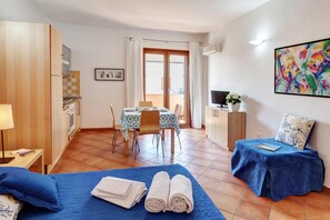 A hotel room with a bed, a dining table with chairs, a kitchenette, a TV, and a balcony with curtains.