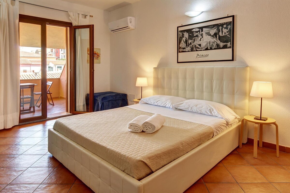 A hotel room with a bed, two bedside lamps, a framed artwork on the wall, and a balcony with a chair.