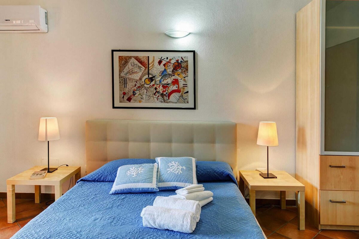 A hotel room with a bed, two bedside tables with lamps, a headboard, a framed abstract painting, and a wardrobe.