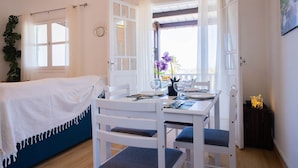 A room with a bed covered in a white blanket, a dining table set with plates and glasses, and a window with curtains.