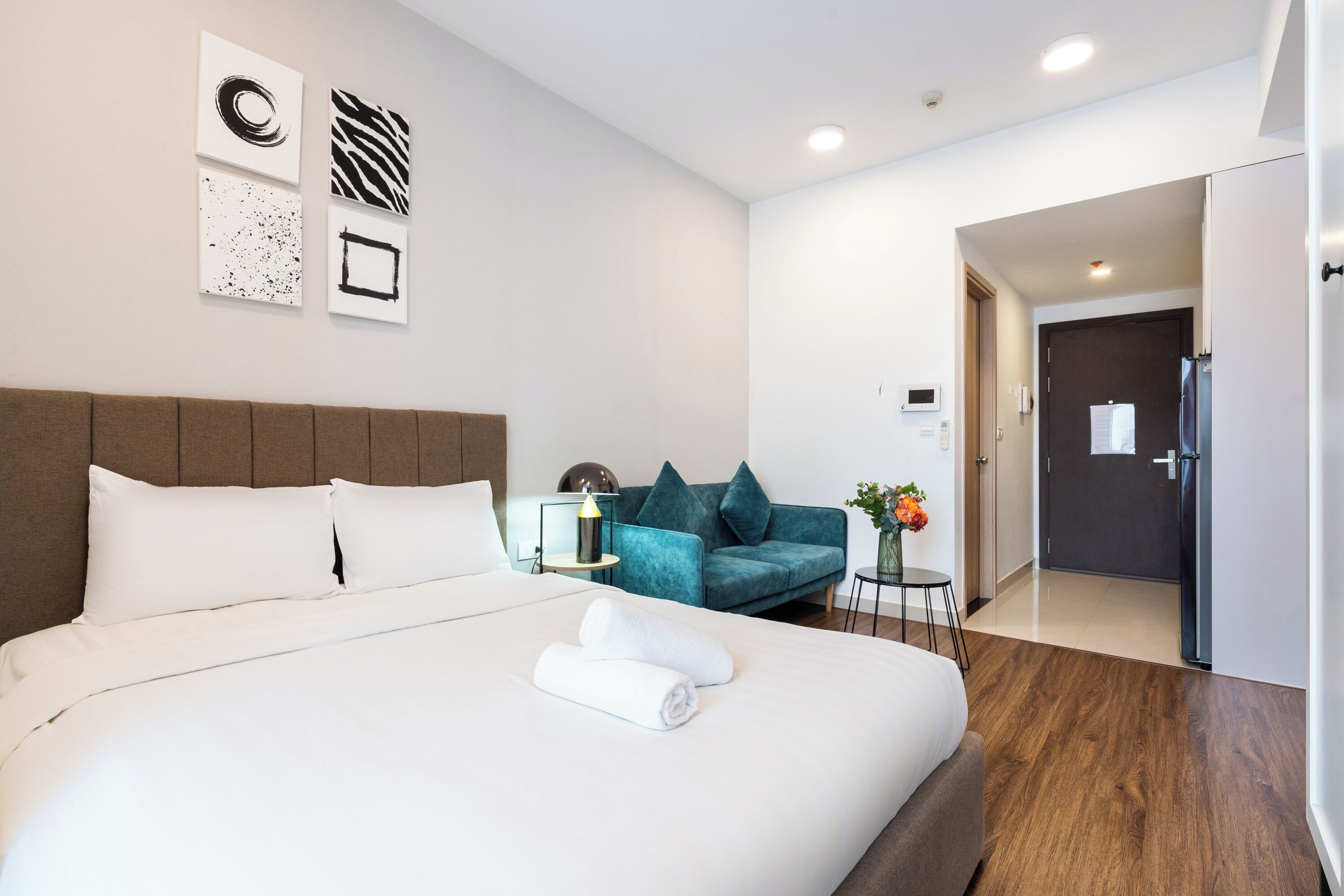 A modern hotel room with a large bed, a teal sofa, and a small side table with a lamp.