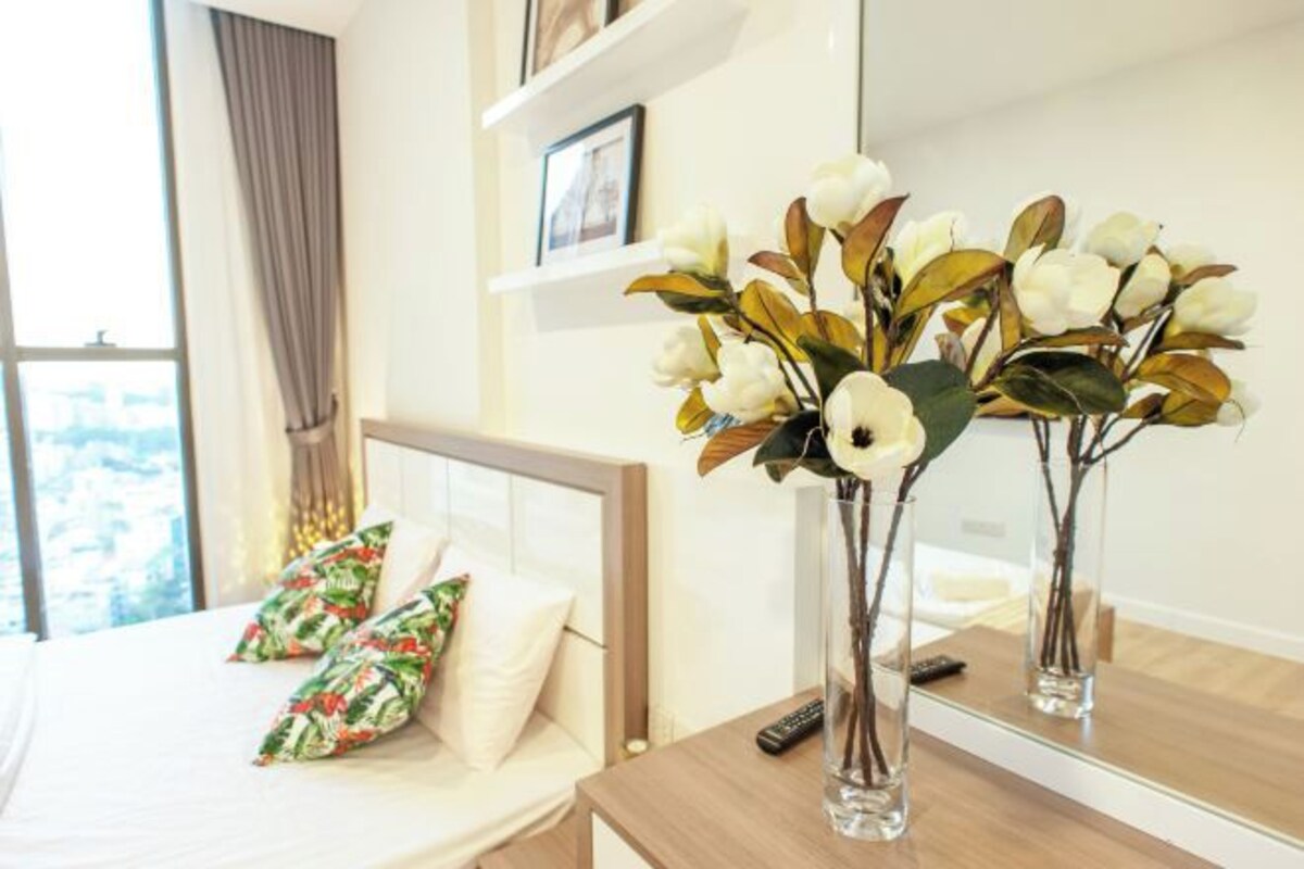 A hotel room with a bed, pillows, a vase with flowers, a remote control, and a mirror reflecting the room.