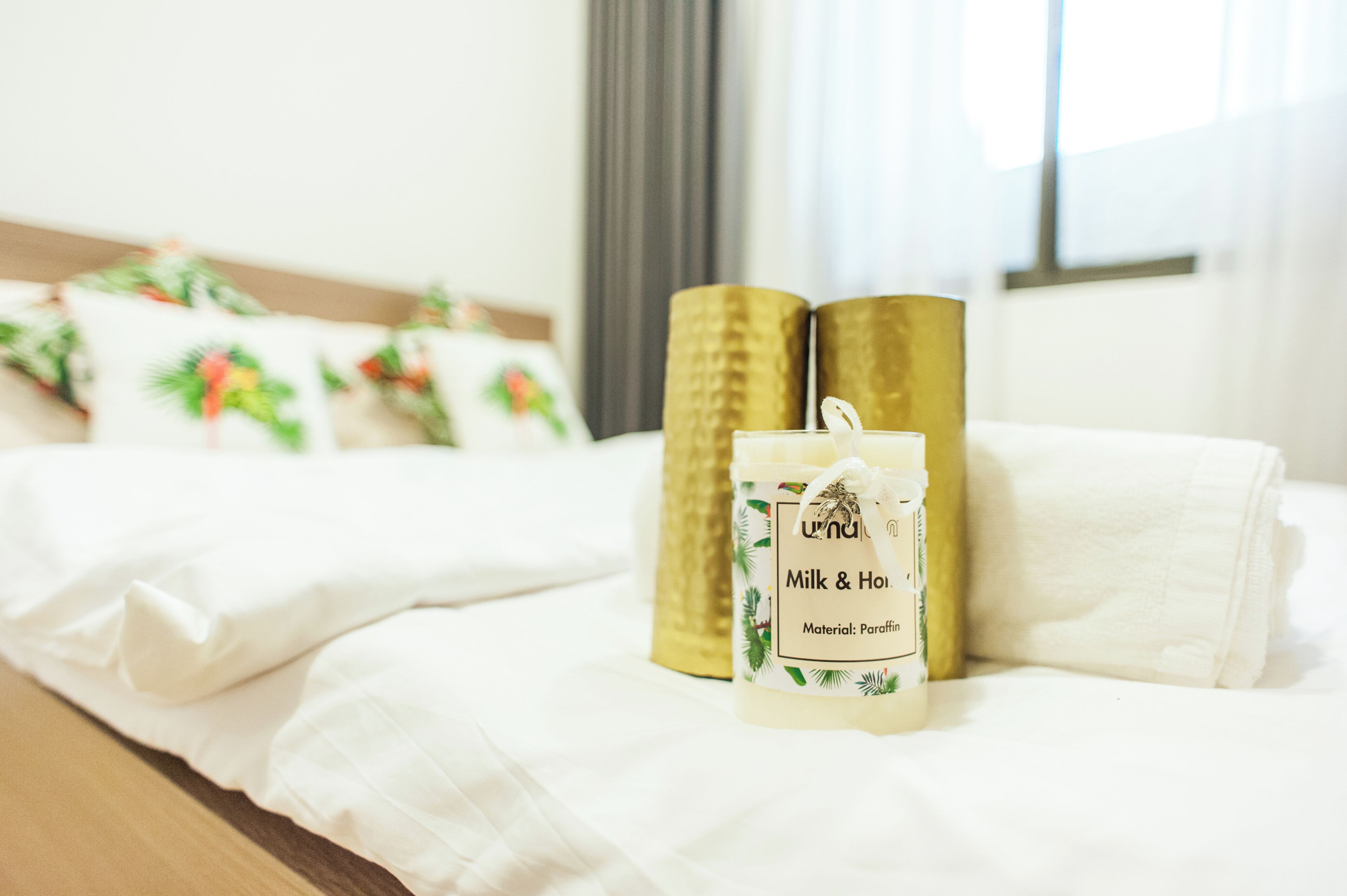 A neatly made bed with white linens, a set of golden pillows, and a packaged candle labeled "Milk & Honey."