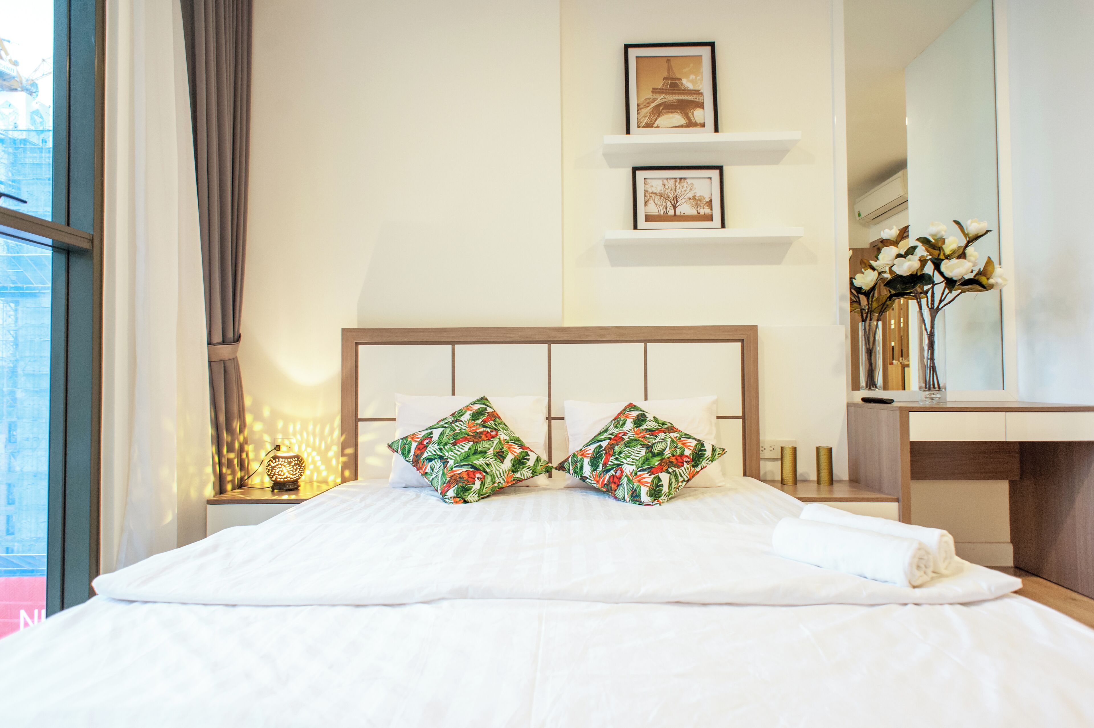 A neatly made bed with white linens and tropical patterned pillows, a bedside table with a lamp, and a wall-mounted shelf with framed pictures.
