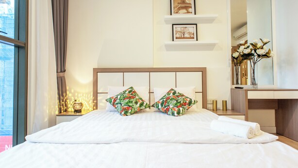 Un lit bien fait, avec du linge de lit blanc et des coussins aux motifs tropicaux, une table de chevet avec une lampe et une étagère murale ornée de tableaux encadrés.