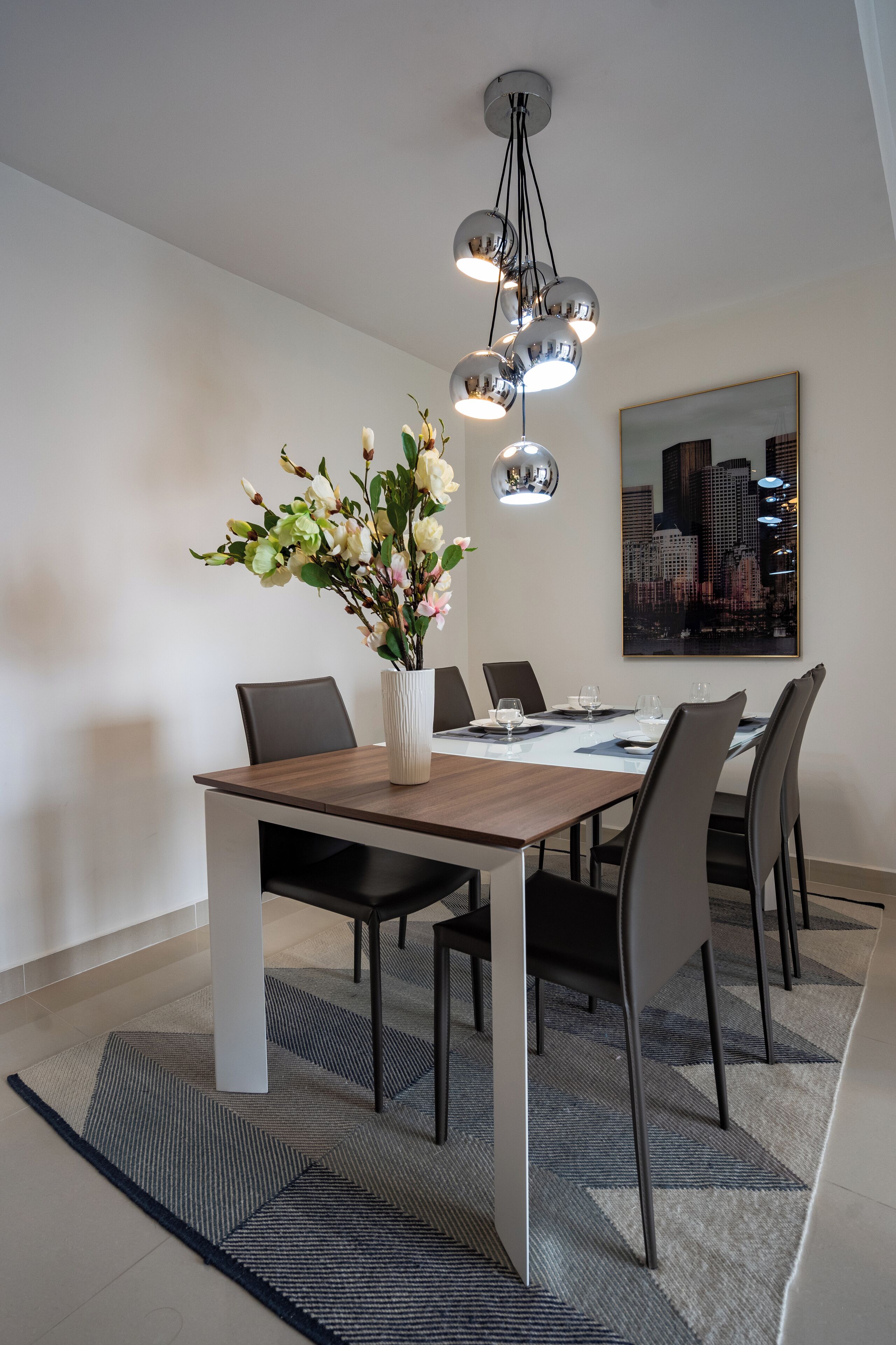 Un comedor moderno con una mesa de madera, sillas negras, un jarrón de flores y un cuadro de un paisaje urbano en la pared.