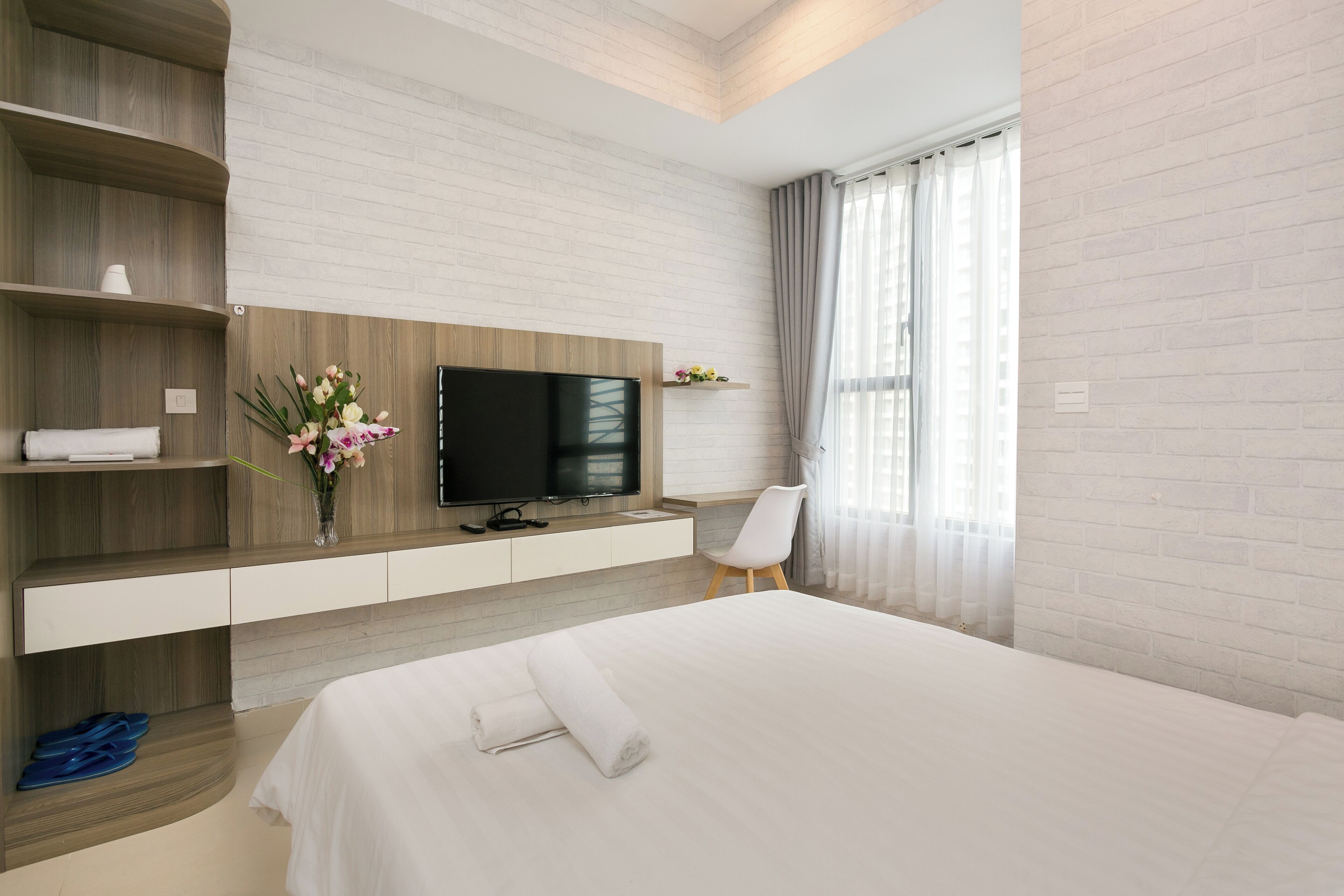 A modern hotel room with a large bed, a flat-screen TV, wooden shelving, a white chair, and a window with curtains.