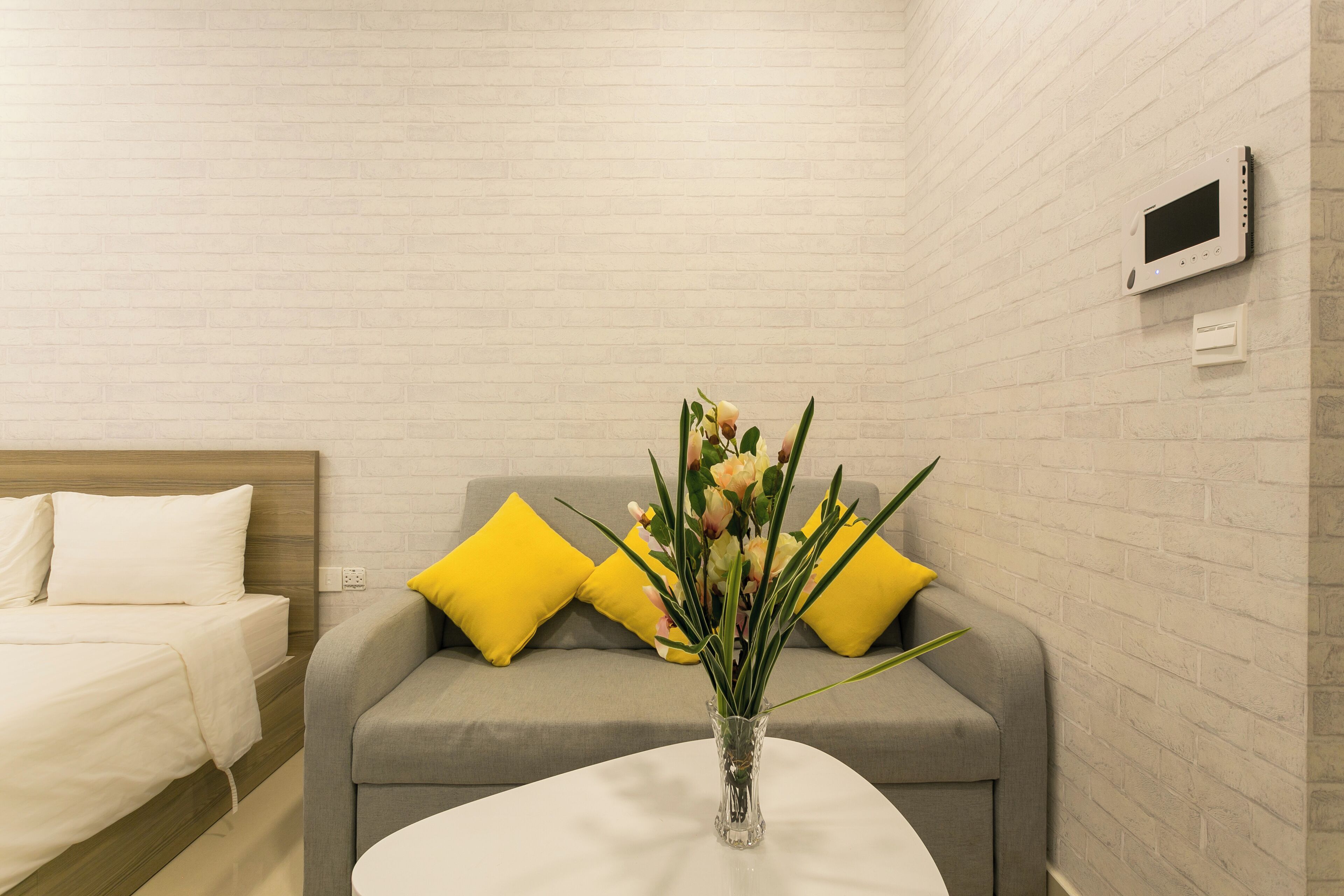 A hotel room with a bed, a gray sofa with yellow pillows, a white round table, and a vase with flowers.