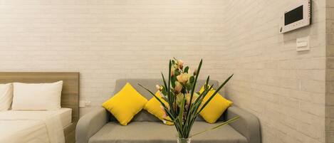 A hotel room with a bed, a gray sofa with yellow pillows, a white round table, and a vase with flowers.