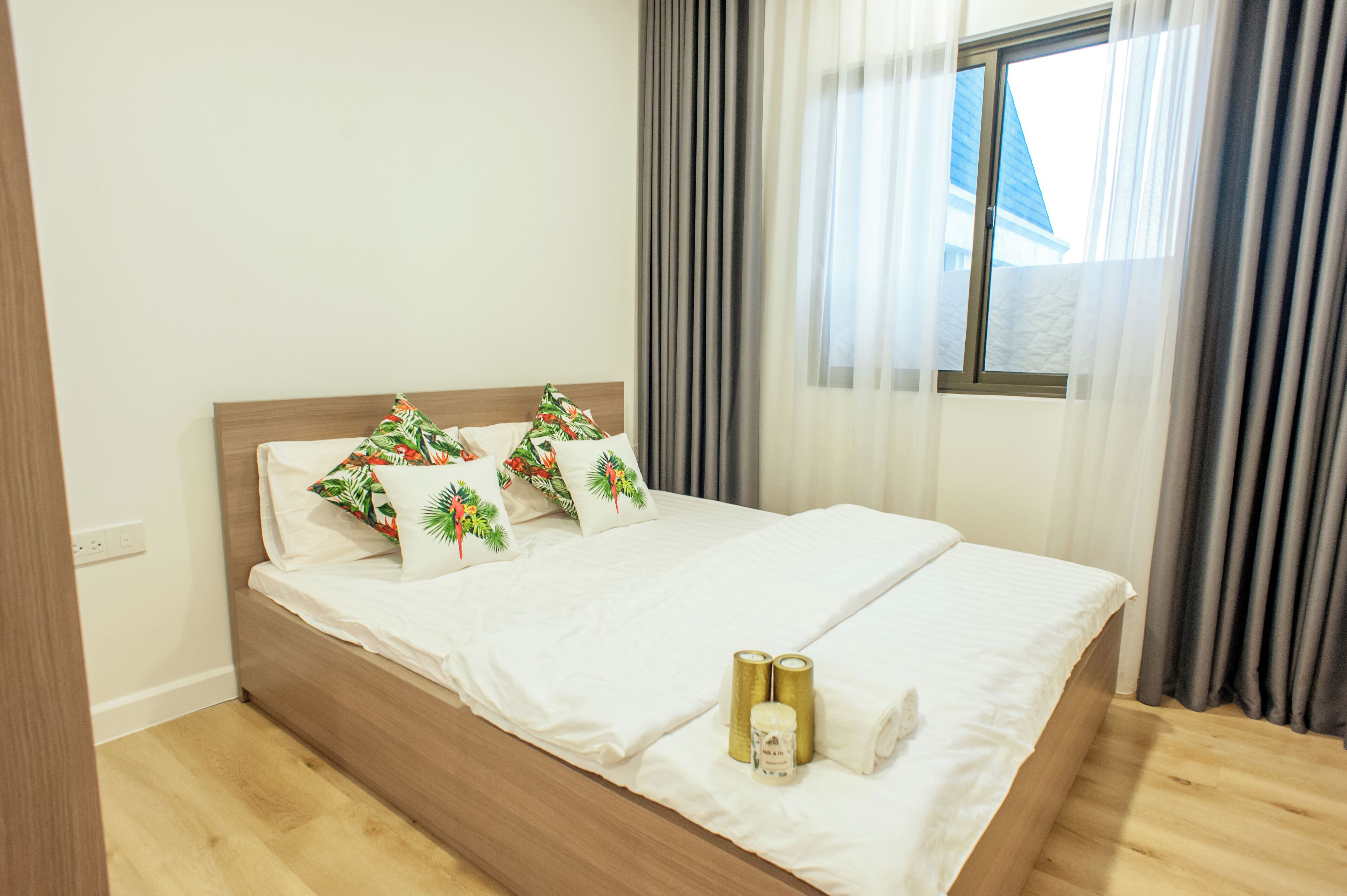 Un lit bien fait, avec du linge de lit blanc et des coussins décoratifs, une tête de lit en bois et une fenêtre avec des rideaux.