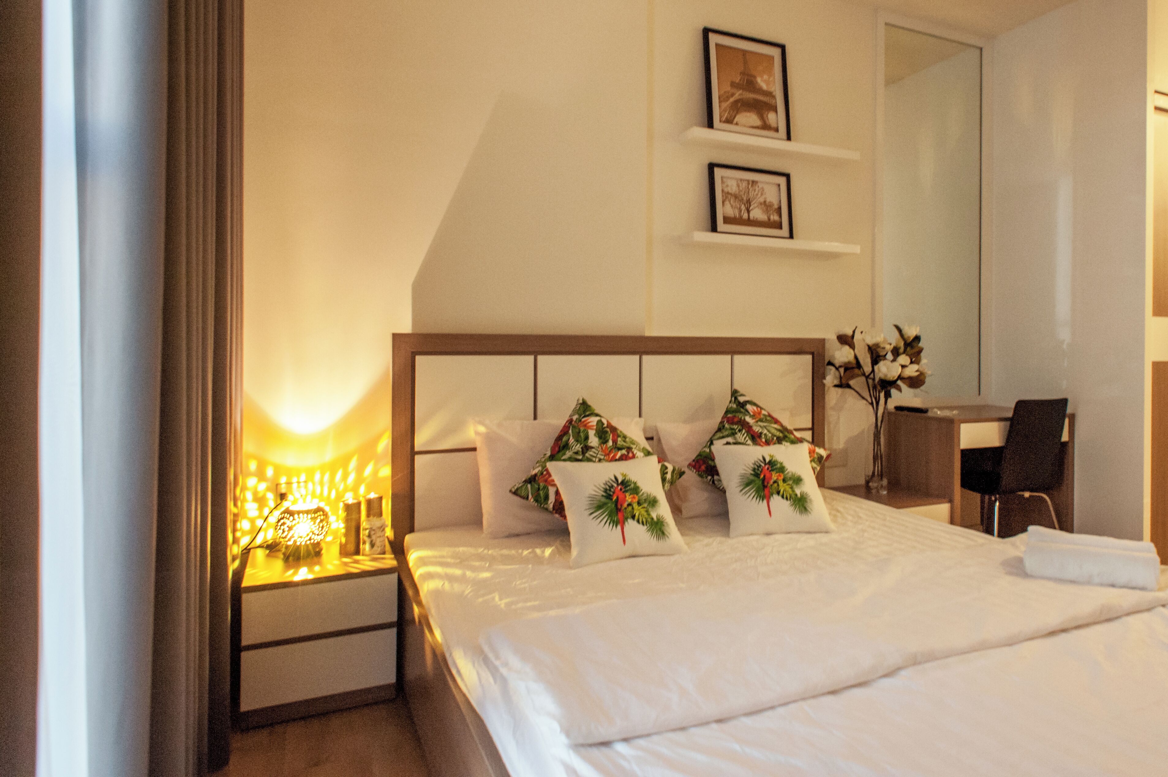 A hotel room with a neatly made bed, a bedside table with a lamp, a desk with a chair, and a mirror on the wall.