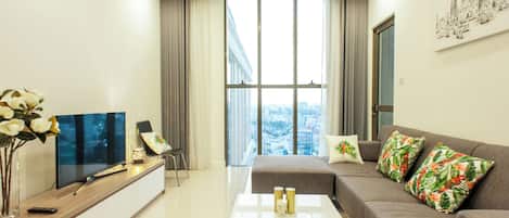 A modern living room with a flat-screen TV, a sofa with tropical pillows, a coffee table, and a large window offering a city view.
