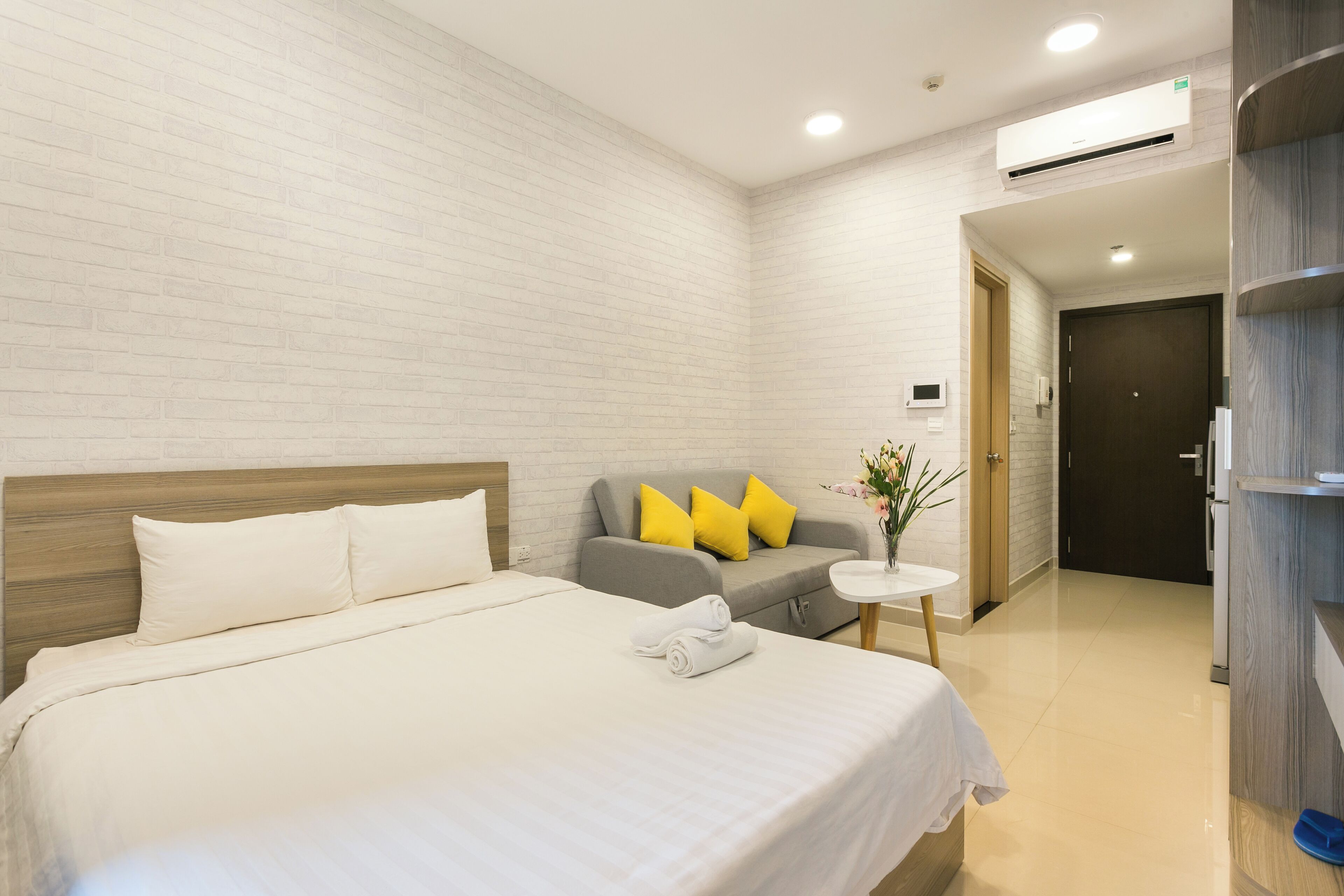 A hotel room with a large bed, a grey sofa with yellow pillows, a small round table with a vase, and a wooden wardrobe.