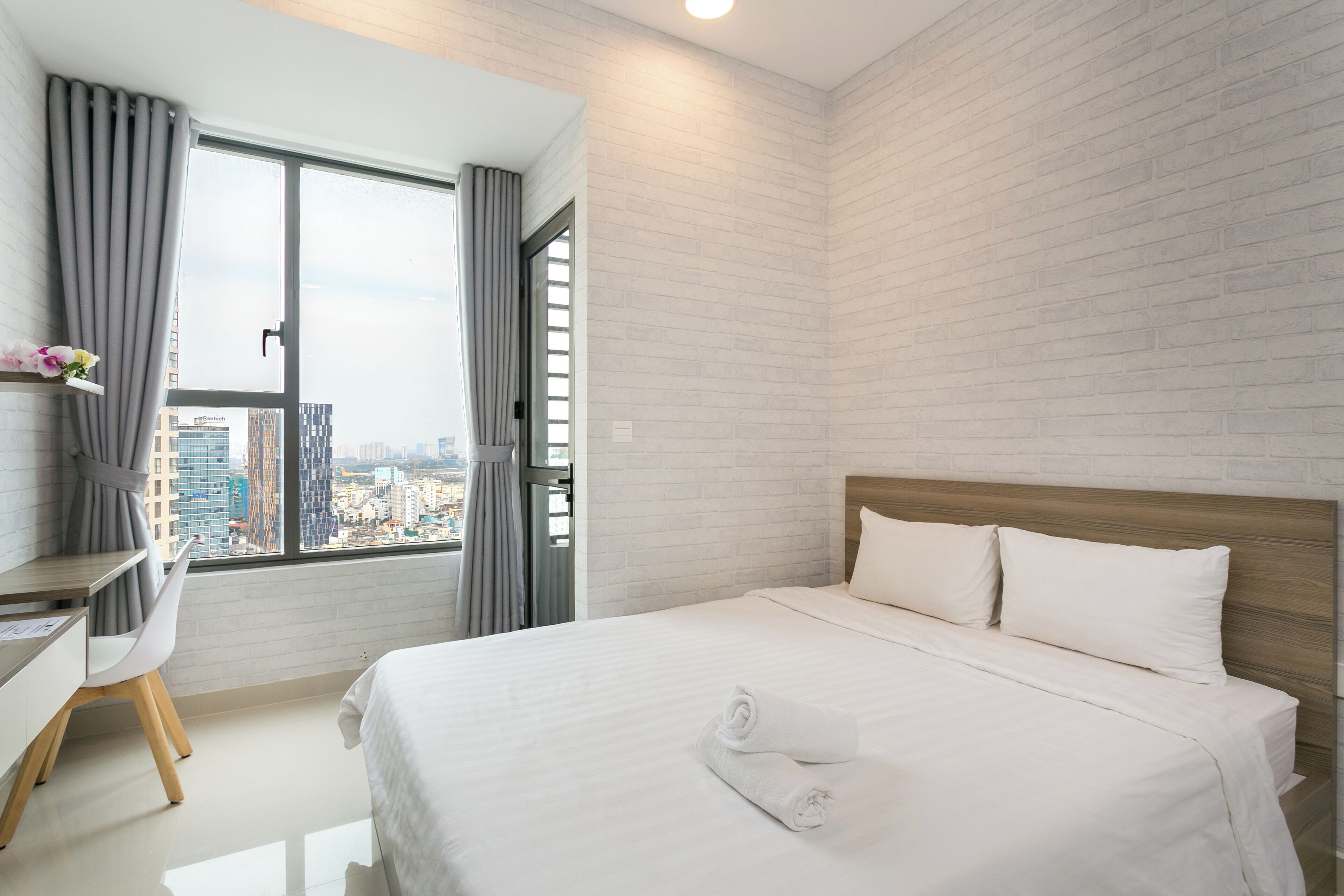 A modern hotel room with a large bed, a desk, and a view of the city through a large window.