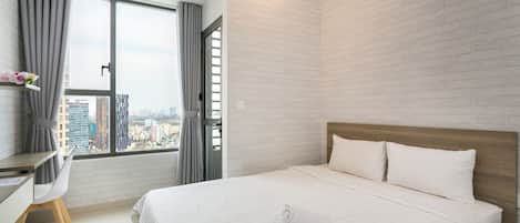 A modern hotel room with a large bed, a desk, and a view of the city through a large window.
